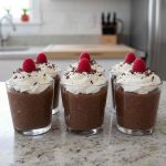 Six keto chocolate chia pudding parfaits topped with whipped cream, chocolate shavings, and a single raspberry, served in clear cups.