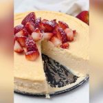 A creamy keto yogurt cheesecake topped with chopped strawberries, with one slice removed to show the soft texture.